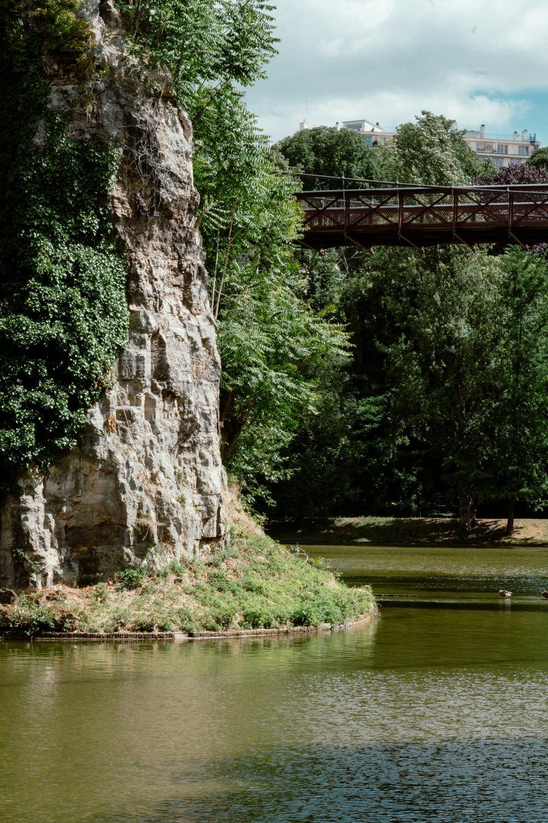 Rocher, lac et passerelle dans un parc parisien vu depuis une allée